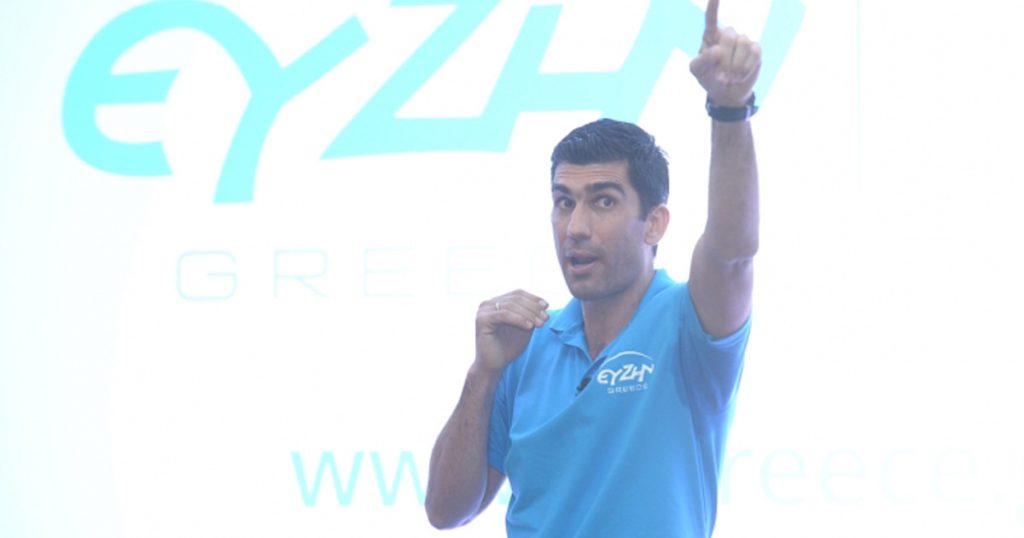 Motivational speaker in a blue polo shirt pointing upward during a seminar, with the EY ZHN Greece logo projected behind him.