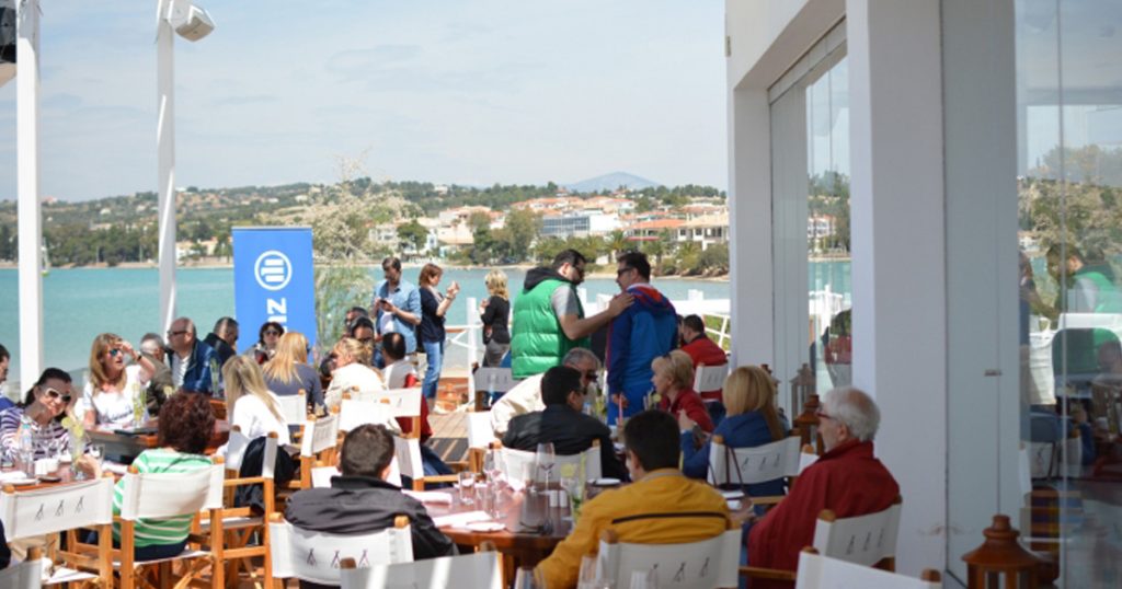 Group of people gathered on a sunny seaside terrace in Porto Heli during a corporate event by Allianz Hellas, with views of the coastal town and turquoise waters in the background.