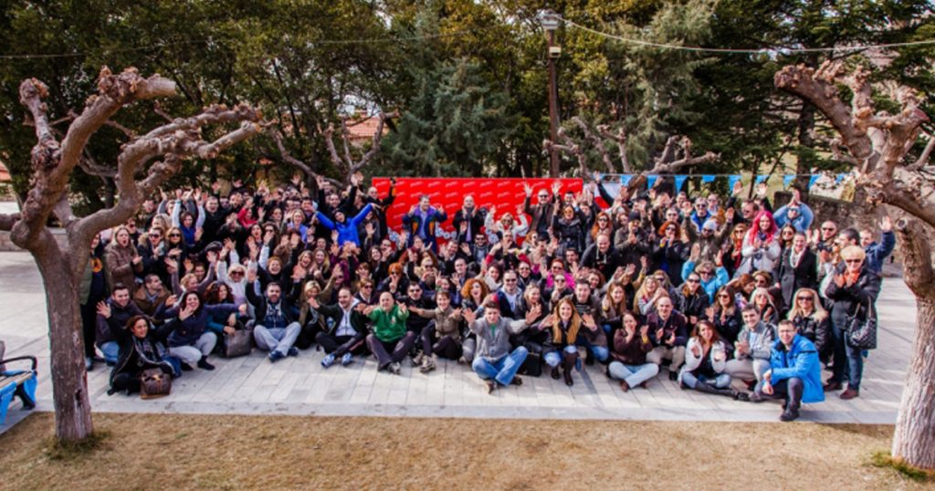 Large group of people posing and cheering outdoors during a corporate treasure hunt event in Arachova, surrounded by leafless trees and a festive backdrop.