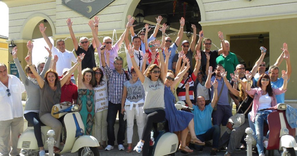 Group of smiling people posing with raised hands in front of a Mediterranean-style building, some sitting on vintage scooters, during AXA Insurance's incentive trip organized by ευΖΗΝ feelόsophy.