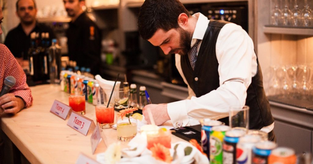 Man dressed in a vest concentrating while mixing cocktails at a bar counter during a team bartending event in Athens, with drinks, mixers, and team signs displayed.