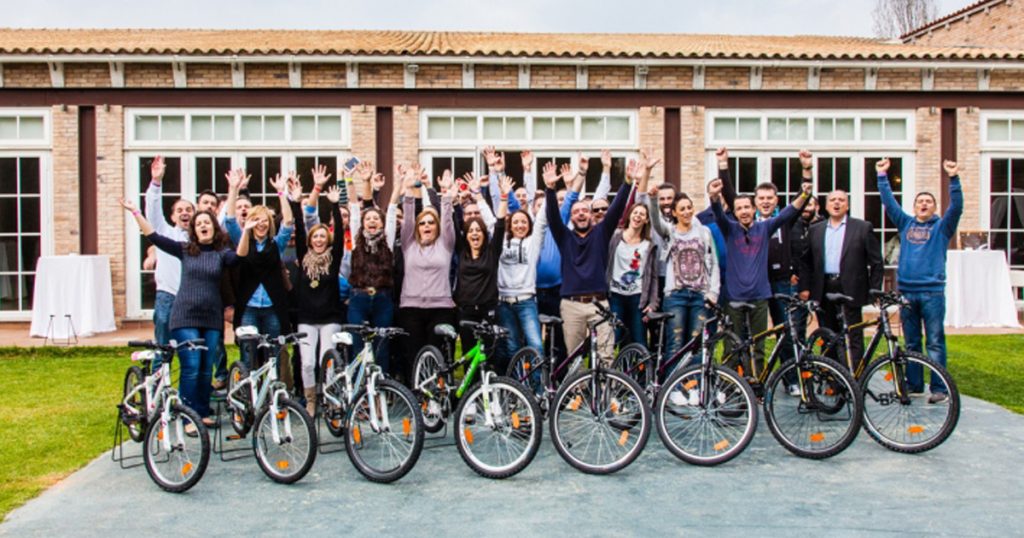 Group of people cheering behind a row of bicycles they assembled during a CSR team-building event by OTE, in front of a building with large windows.