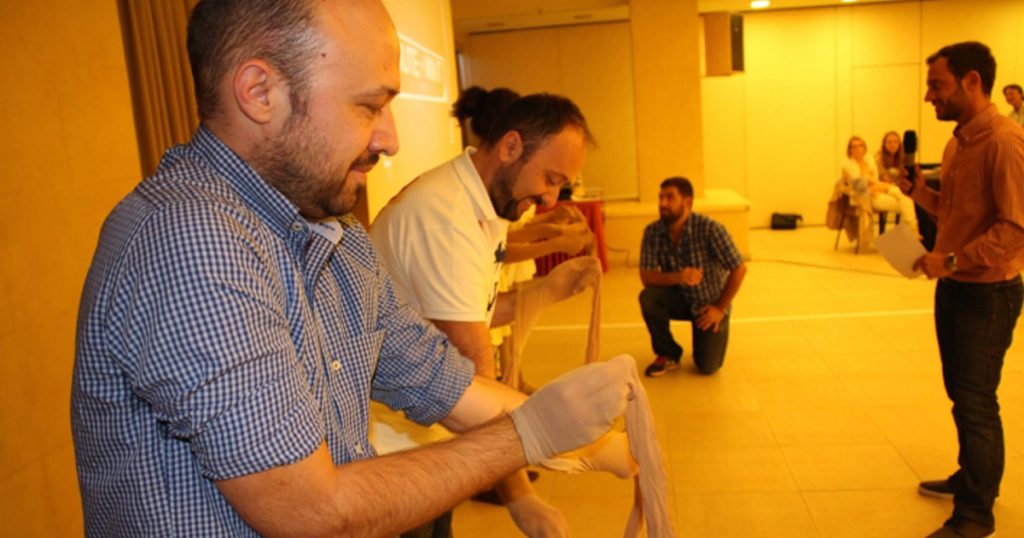 Group of adults participating in a fun indoor team challenge, unrolling bandages as part of a 'Minute to Win It' game, with a host giving instructions.