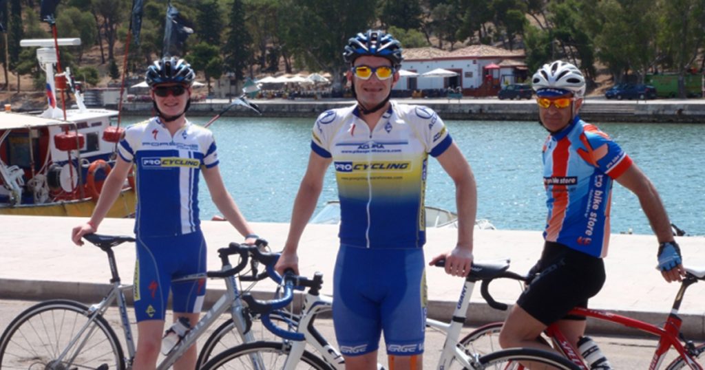Three cyclists in professional gear, including a father and son, posing with their road bikes by the waterfront in Greece, with boats and trees in the background.