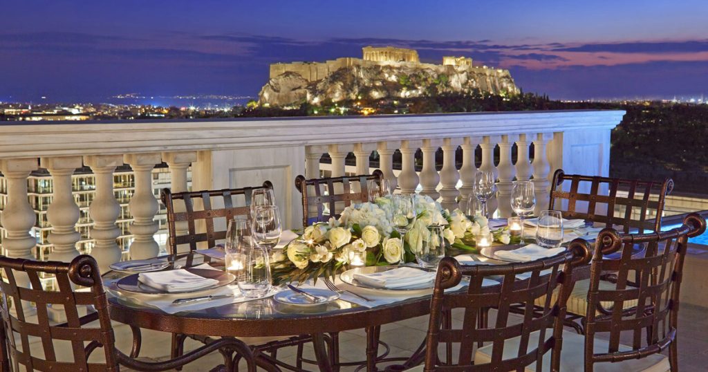 Elegant rooftop dinner setup with candlelight and white floral centerpiece overlooking the illuminated Acropolis of Athens at dusk, representing the essence of luxury experiences in the Greek capital.
