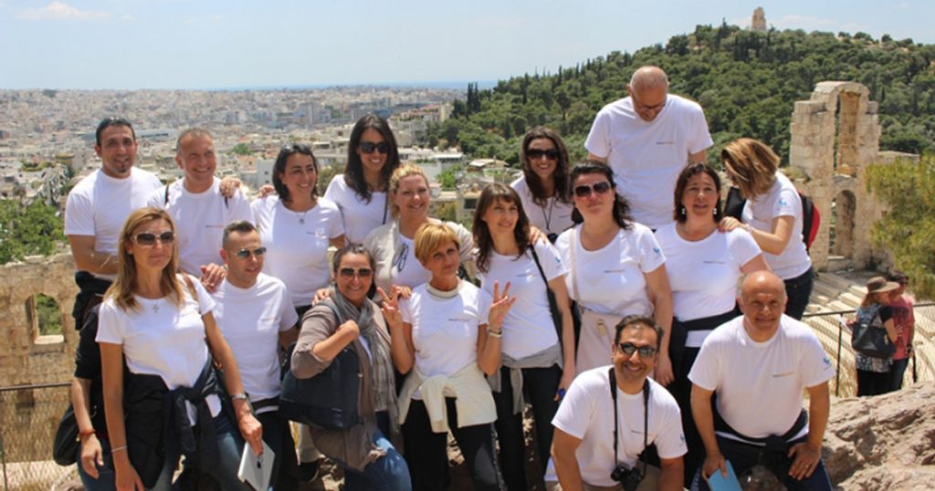 Team members from Novo Nordisk Italy posing for a group photo on a sunny day at the Acropolis in Athens, with the cityscape and ancient ruins in the background.