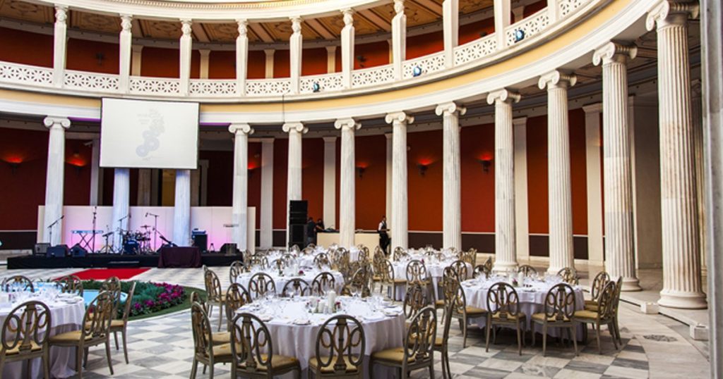 Elegant round tables set for a formal event inside a neoclassical rotunda with tall marble columns and a central stage, during the MIDIS Group annual conference in Athens organized by ευΖΗΝ feelόsophy.