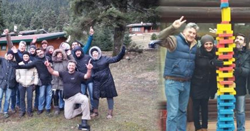 Team of people dressed in winter clothes celebrating outdoors in a snowy forest setting, next to a tall colorful Jenga tower built as part of a team-building challenge.