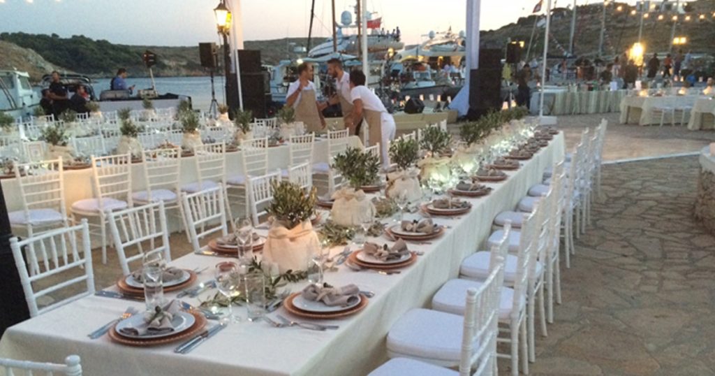 Elegant seaside wedding dinner setup on a Greek island with white chairs, long decorated tables, olive centerpieces, and ambient lighting at sunset.