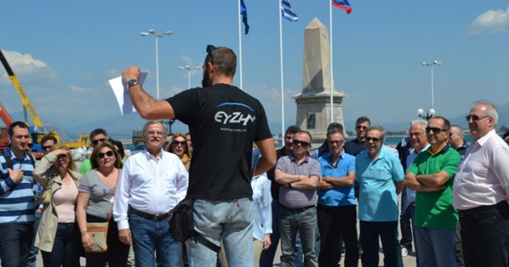 A group of National Bank of Greece employees gathered outdoors, listening to a team-building facilitator wearing a black EY ZHN shirt, with flags and a monument in the background.