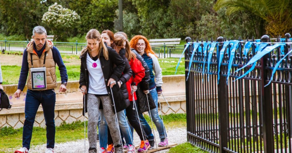 A group of people participating in a team-building activity involving coordination and walking together on wooden planks, during a Nike Hellas outdoor event in a park.