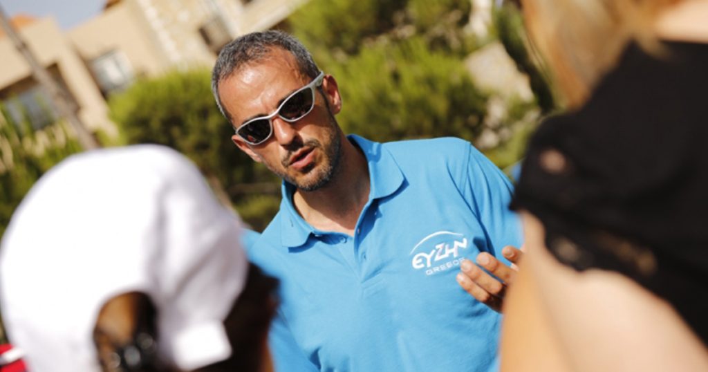 Man wearing sunglasses and a blue 'ευΖΗΝ Greece' polo shirt speaking to a group of people during a corporate meeting at Costa Navarino.