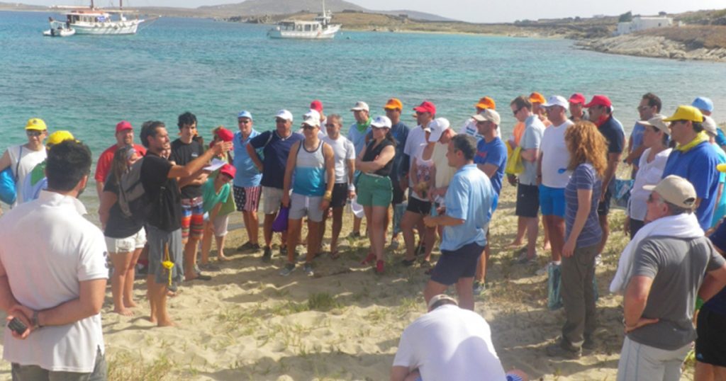 Corporate team bonding activity on a sandy beach at Rinia island near Mykonos, with a large group of participants gathered around a facilitator, wearing casual summer clothing and colorful caps.