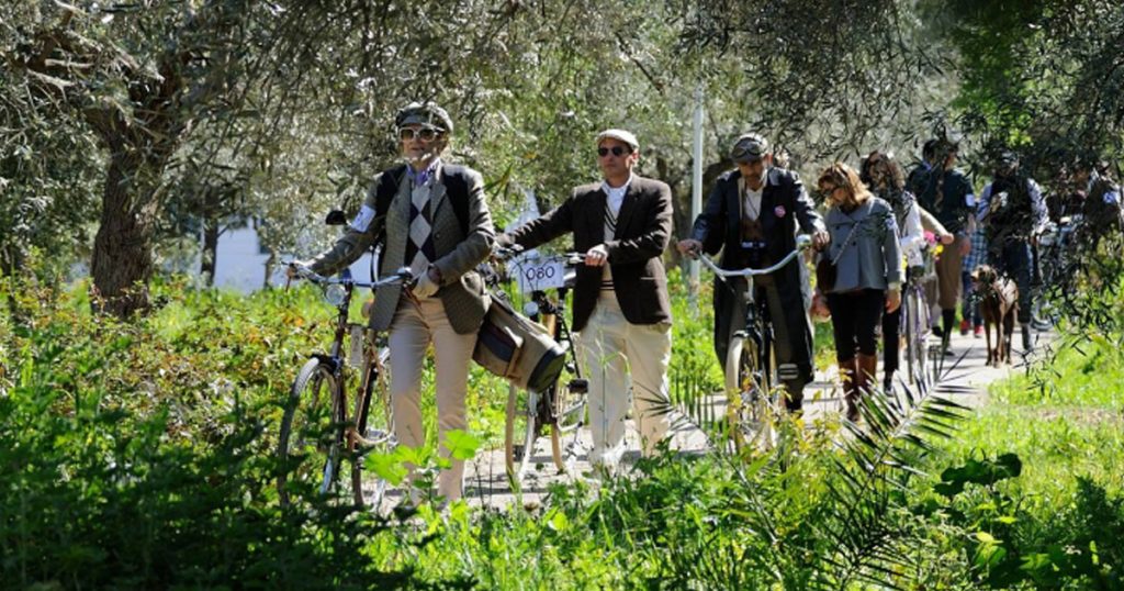 Participants dressed in vintage tweed outfits walking their classic bicycles through a lush olive grove during the 2nd Tweed Run event in Spetses, Greece.