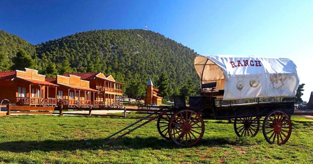 A sunny day at a Western-style ranch with wooden buildings and a vintage covered wagon labeled 'RANCH', set against a backdrop of pine-covered hills—capturing the essence of a cheerful spring outing.