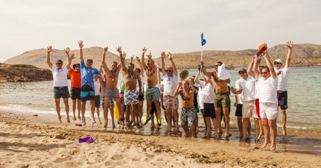 Group of men celebrating on a sandy beach after completing a team-building survival game on a remote Greek island, wearing medals and casual swimwear.