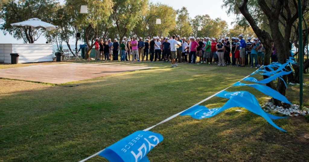Large group of people gathered on a lawn near the sea, participating in outdoor team bonding activities organized by ευΖΗΝ feelόsophy, with branded blue flags marking the area.