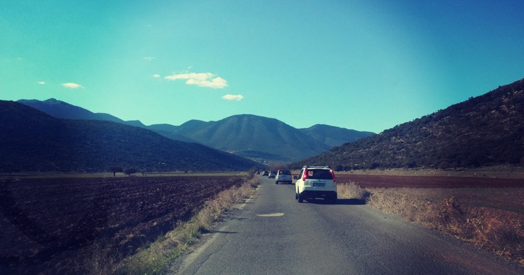 Convoy of cars driving through a scenic rural road surrounded by mountains under a clear blue sky, during a corporate road trip or incentive event.