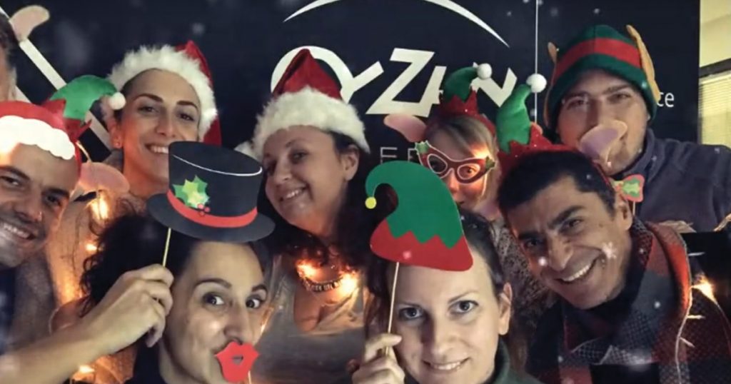 Group of colleagues dressed in festive Christmas accessories and smiling in front of a 'ευΖΗΝ feelόsophy' backdrop, celebrating the holidays together.