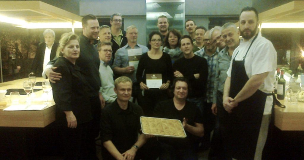 Group of people posing in a modern kitchen after a Greek cooking class, with one person holding a tray of traditional dessert and a chef standing in front.