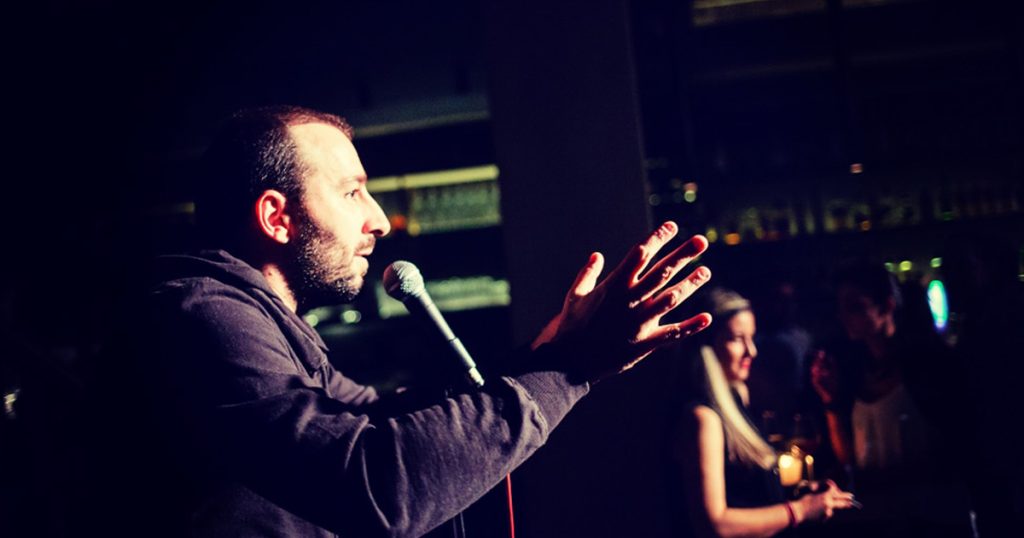 Man performing stand-up comedy with expressive gestures at a lively Christmas corporate event, holding a microphone while an audience enjoys the show in a warmly lit venue.