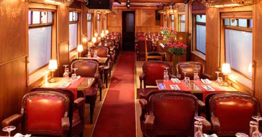 Elegant vintage-style train dining car set up for a theatrical performance in English, with plush red leather chairs, dim table lamps, and wooden paneling creating a warm, immersive atmosphere.