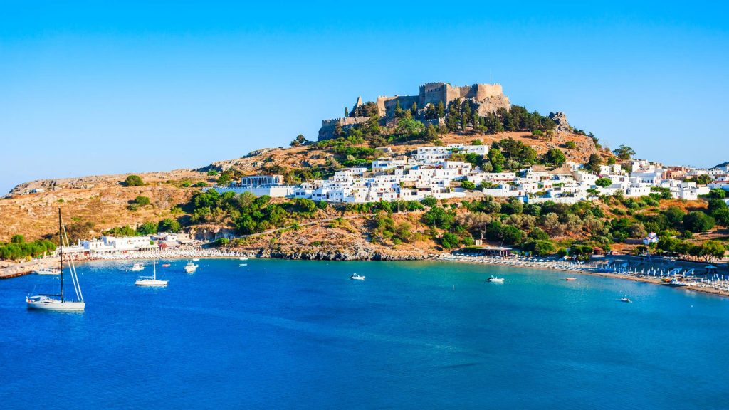 Scenic view of Lindos village and ancient acropolis in Rhodes, Greece, with yachts anchored in the turquoise bay.
