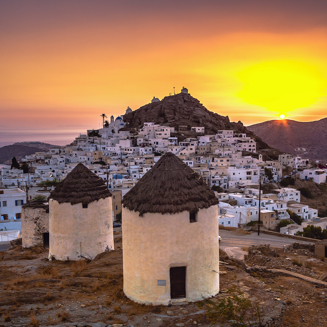 Traditional windmills overlooking Chora in Ios, Greece, at sunset with whitewashed houses and golden sky
