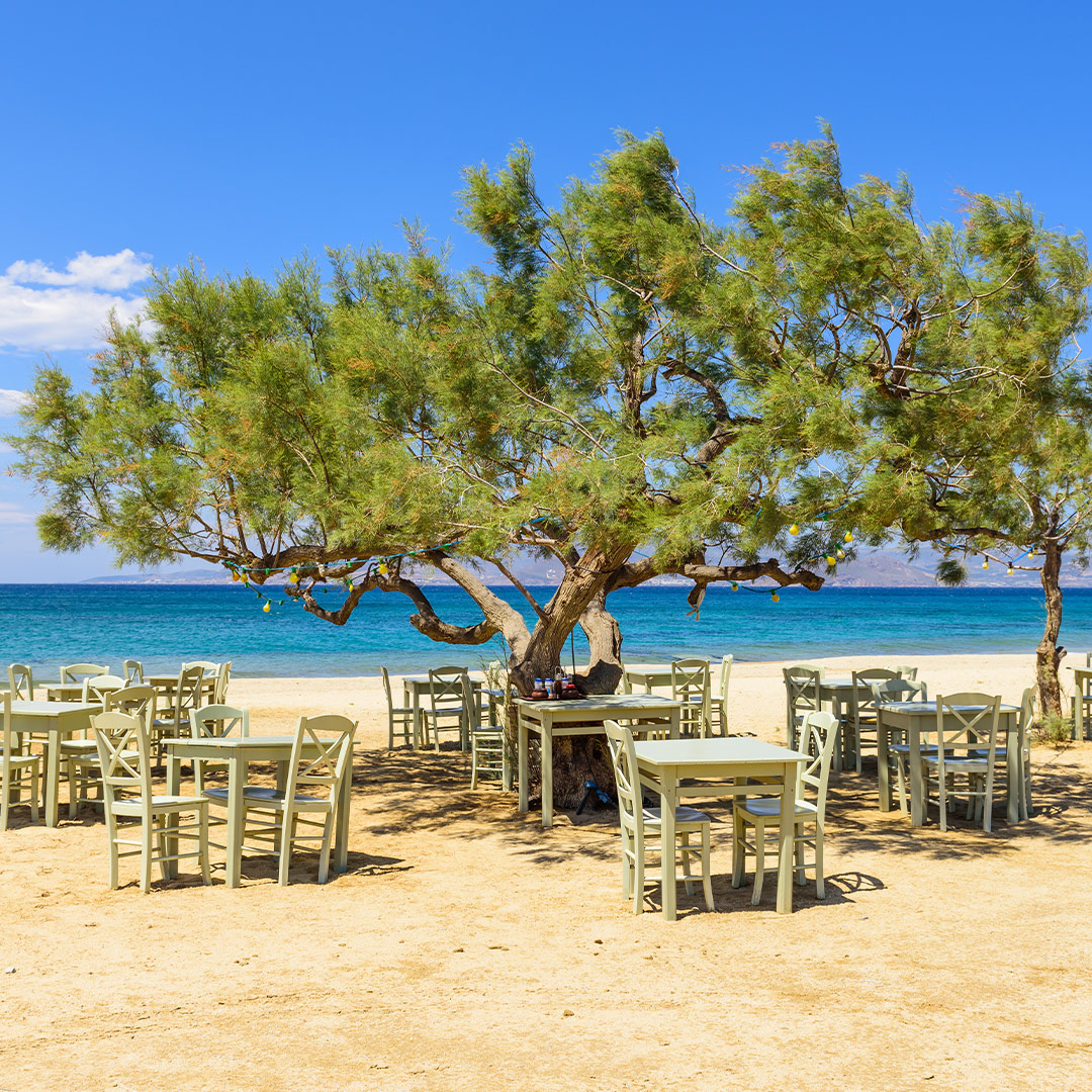 Seaside taverna tables under a tree on a sandy beach in Naxos, Greece, with turquoise waters and clear blue sky.