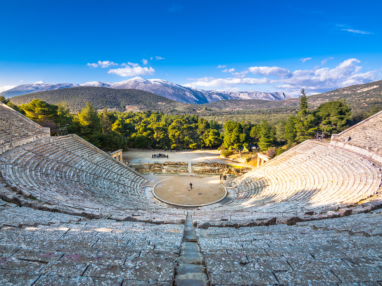 Panoramic view of the ancient Epidaurus theater in Greece, with stone seats, lush forest, and mountain backdrop.
