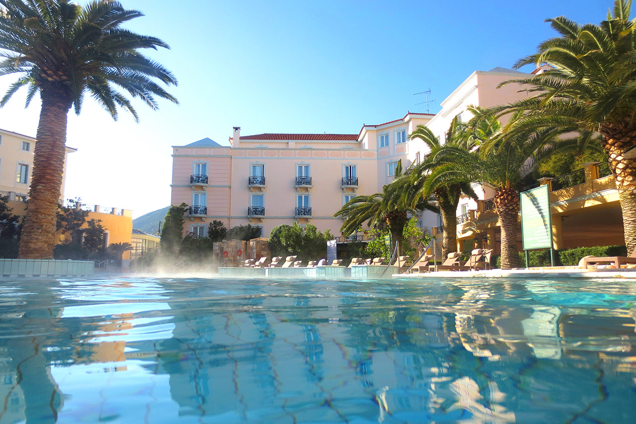 Outdoor thermal pool surrounded by palm trees and a neoclassical spa building under a clear blue sky in Greece.