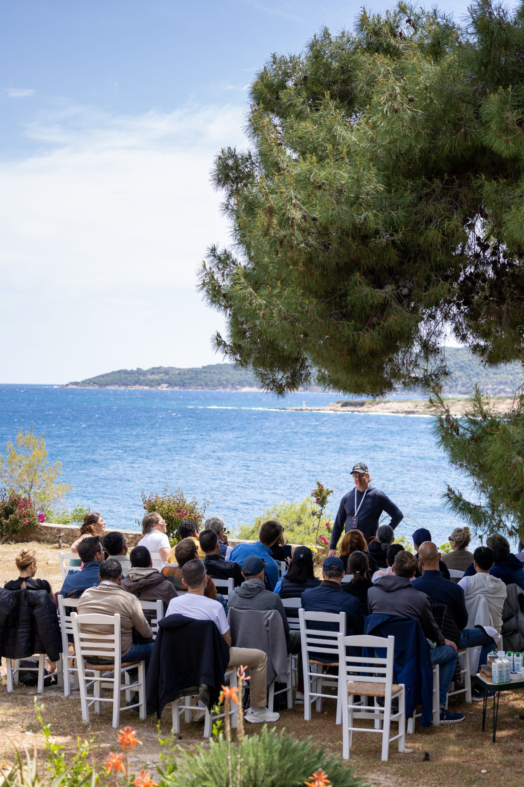 Outdoor keynote speaker addressing seated audience by the sea in Spetses, with blue waters and pine trees creating a serene setting.